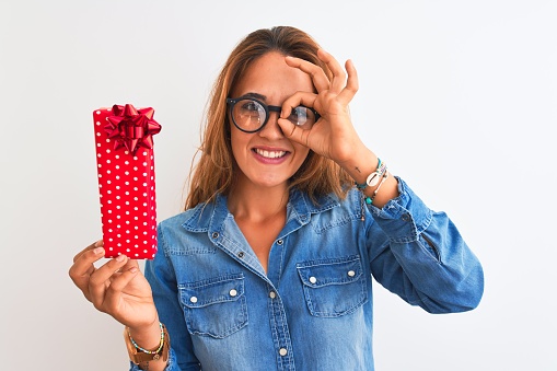 Young beautiful redhead woman wearing glasses and holding birthday present with happy face smiling doing ok sign with hand on eye looking through fingers
