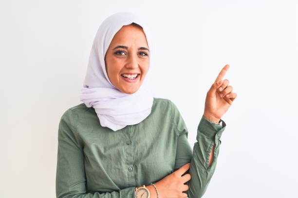 joven hermosa mujer árabe con hiyab musulmán tradicional sobre fondo aislado con una gran sonrisa en la cara, apuntando con la mano y el dedo hacia un lado mirando a la cámara. - women open traditional culture human hand fotografías e imágenes de stock