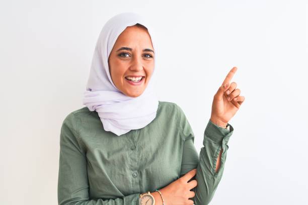 고립된 배경 위에 전통적인 이슬람 히잡을 입은 젊은 아름다운 아랍 여성은 얼굴에 큰 미소를 지으며 카메라를 바라보는 측면을 손으로 손가락으로 가리킵니다. - women open traditional culture human hand 뉴스 사진 이미지