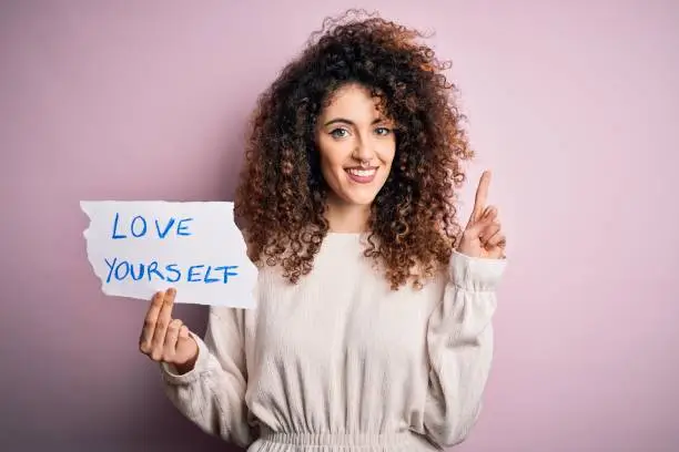 Young beautiful woman with curly hair and piercing holding paper with love yourself message surprised with an idea or question pointing finger with happy face, number one