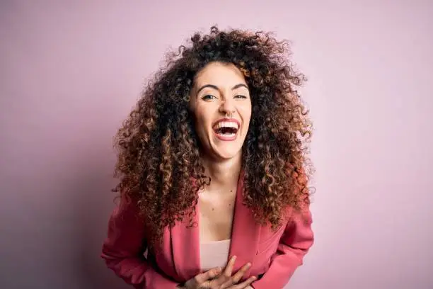 Young beautiful businesswoman with curly hair and piercing wearing elegant jacket smiling and laughing hard out loud because funny crazy joke with hands on body.