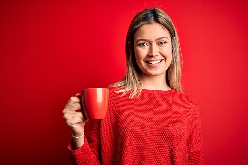 Young beautiful woman drinking cup of coffee standing over isolated red background with a happy face standing and smiling with a confident smile showing teeth