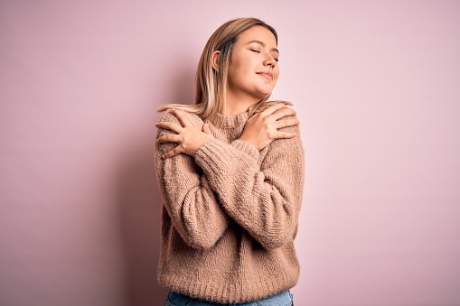 Young beautiful blonde woman wearing winter wool sweater over pink isolated background Hugging oneself happy and positive, smiling confident. Self love and self care