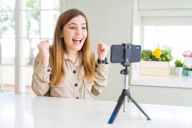 istock Beautiful young woman doing online video call using smartphone webcam celebrating surprised and amazed for success with arms raised and open eyes. Winner concept. 1205037135