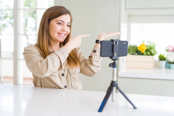 istock Beautiful young woman doing online video call using smartphone webcam amazed and smiling to the camera while presenting with hand and pointing with finger. 1205037206