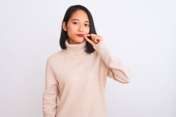 junge chinesin trägt rollkragenpullover, der über einem isolierten weißen hintergrundmund und lippen wie reißverschluss mit denfingern geschlossen ist. geheim und still, tabu-gespräche - finger in mouth women turtleneck serious stock-fotos und bilder