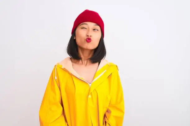 Photo of Young beautiful chinese woman wearing raincoat and wool cap over isolated white background making fish face with lips, crazy and comical gesture. Funny expression.