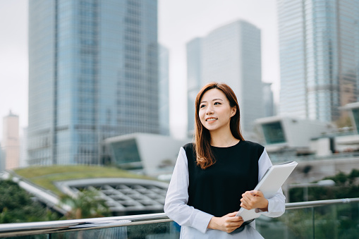 Portrait of confidence young Asian businesswoman holding paperwork and laptop against city scene in front of modern office buildings