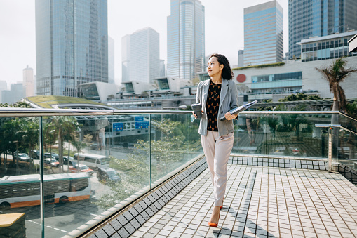Portrait of confidence Asian businesswoman looking away and holding documents and having coffee to go against city scene in front of modern office buildings
