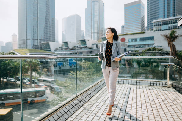自信的亞洲女商人在現代辦公樓前望去，拿著檔，喝咖啡，與城市場景相對抗 - choicesea 個照片及圖片檔