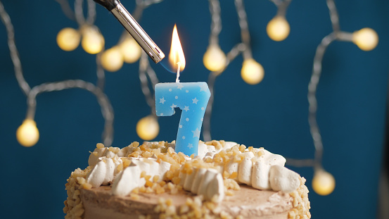 Birthday cake with 7 number candle on blue backgraund set on fire by lighter. Close-up view