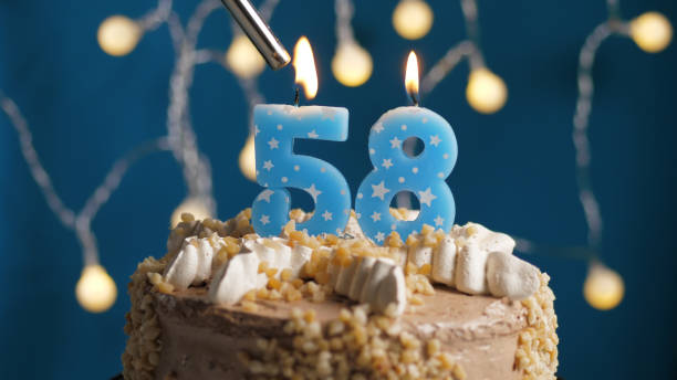 istock Birthday cake with 58 number candle on blue backgraund set on fire by lighter. Close-up 1205065788