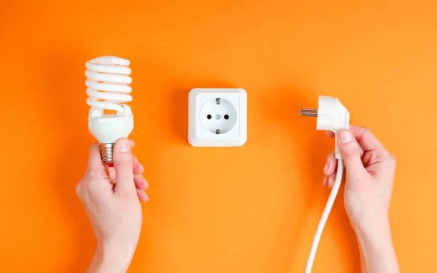 istock Female hands plug the power plug into electrical outlet while holding spiral light bulb on orange background. Minimalism 1205080899
