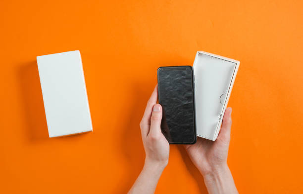 mani femminili che tengono nuovo smartphone nella scatola. unboxing vista dall'alto su sfondo arancione, minimalismo - unboxing foto e immagini stock
