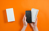Female hands holding new smartphone in the box. Unboxing Top view on orange background, minimalism