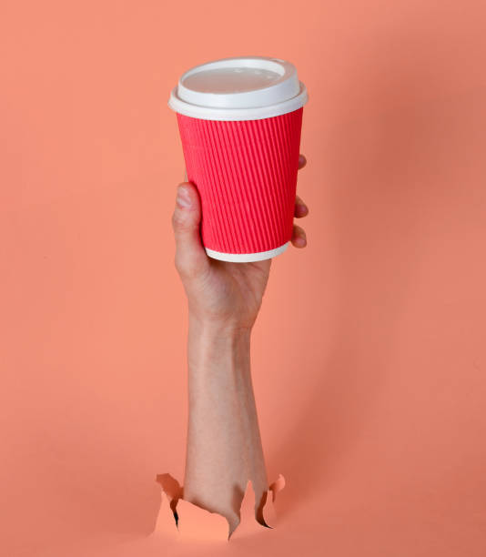 Female hand holds paper coffee cup through torn pink paper. Minimalistic concept