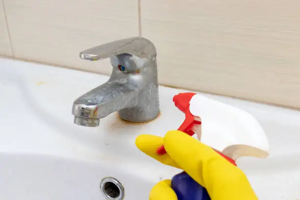 Hand in yellow rubber gloves holding a spray liquid cleaning agent for washing dirty faucet with limescale, calcified water tap with lime scale on washbowl in bathroom, house cleaning concept.