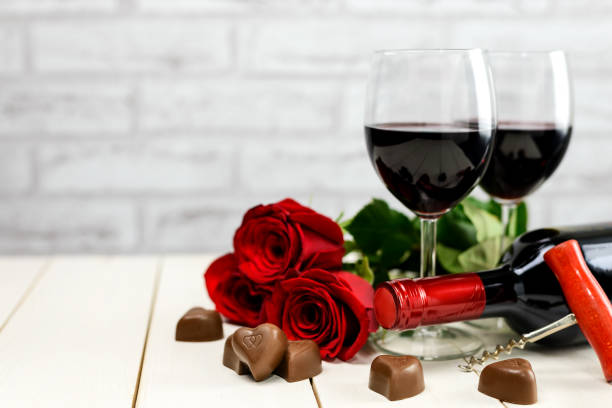 A glasses of wine, wine bottle, roses and chocolate hearts Valentine's Day concept. Two glasses of wine, wine bottle, corkscrew, red roses and chocolate hearts on a white wooden table with copy space for text. Selective focus. rose bouquet red table stock pictures, royalty-free photos & images