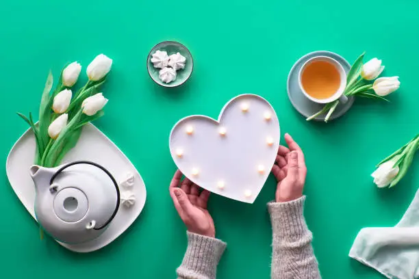 Spring geometric flat lay. Female hands show heart shape sign. Tea cup, tea pot, sweets and white tulips on green table. Mothers day, international women day 8 March or your mom's birthday.