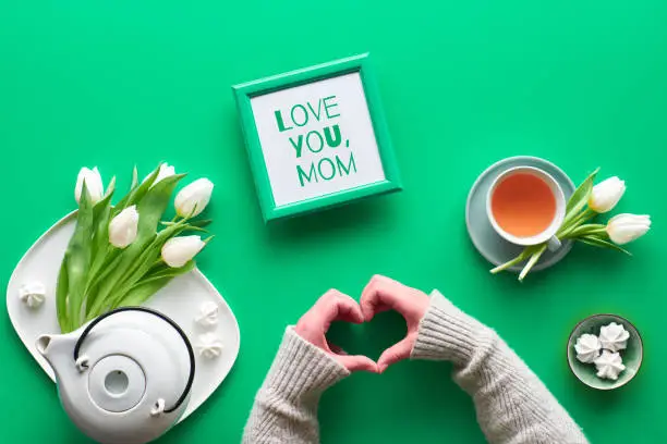 Spring celebration flat lay. Female hands show heart shape sign. Tea cup, tea pot, sweets and white tulips on green table. Mothers day, international women day 8 March or your mom birthday.