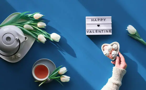 Happy Valentine day celebration flat lay. Female hands show heart shape sign. Tea cup, tea pot, sweets and white tulips on tale, background in trendy classic blue color.