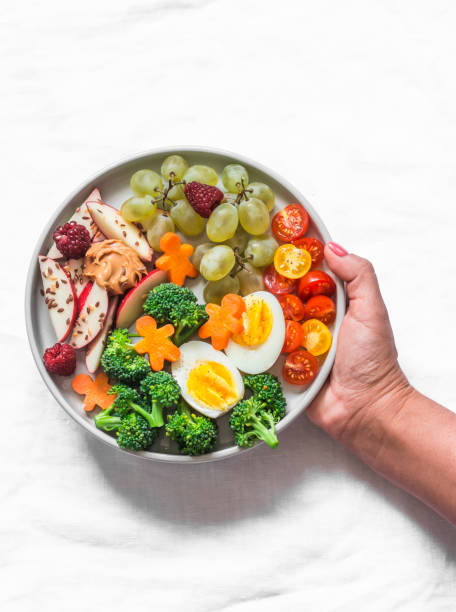 istock Women hand with a large snack plate. Boiled egg, broccoli, carrots, apples with peanut butter, grapes - delicious diet breakfast, snack on a light background, top view 1205177270