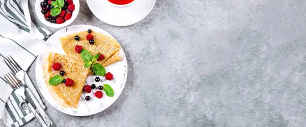 Delicious Crepes Breakfast on gray concrete table background. Orthodox holiday Maslenitsa. Pancakes with berry black currant, raspberry, jar of honey and mint. White cup of tea. Top view, copy space, banner for website