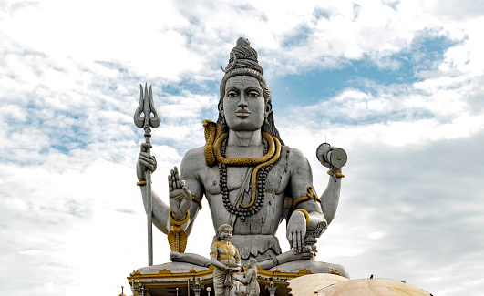 Very Famous and among the tallest Statue of Lord Shiva, meditating in Lotus Pose with Trident, drum in hand, snake around neck while giving blessings to His followers. This photo has a dramatic sky background adding to the glory of Lord Shiva. Lord Shiva is Hindu mythology god and is worshiped by yogis of India and all over the world. This famous statue is situated in the town of Murudeshwara, Karnataka India, and has many folklores associated with it.
