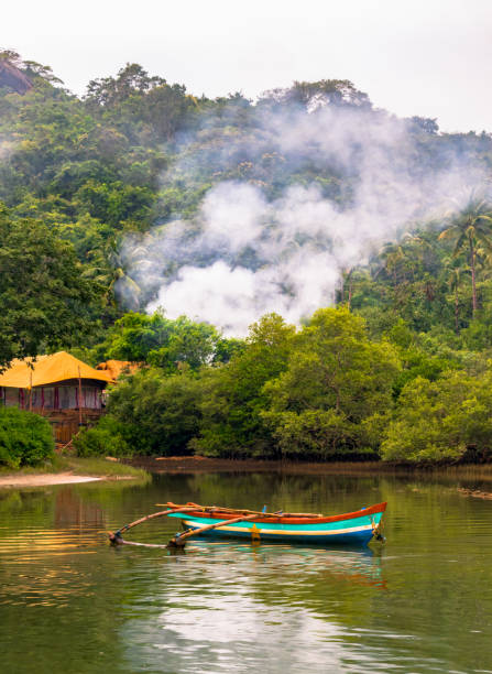 Mesmerizing tropical landscape of remote coastal area comprising mangrove forest with hill and clouds, a temporary cottage for fishing enthusiasts and a wooden canoe for rowing and fishing in the pond Vertical photo of a beautiful scenery of a camping site amidst lush green forests of Western ghats with a lake in foreground. Smoke from the bonfire could be seen in the photo, and there is a small colorful canoe floating in the lake for fishing or for leisure. As is evident from the photo, it is an ideal camping site for adventure lovers and thrill seekers. It is very close in the lap of nature, far away from the hustle bustle of a daily city life. palolem beach stock pictures, royalty-free photos & images
