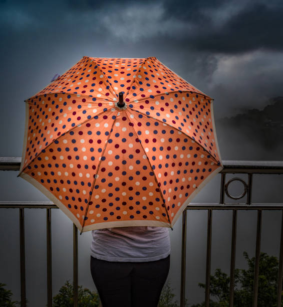 Beautiful photo depicting the mesmerizing scene of a rainy season downpour. An unrecognizable girl in early 20s is holding a polka dot umbrella and is gazing the clouds crossing her covering all the sunlight. The colorful umbrella she is holding shows her fashion sense and that she is trendy, independent and a lone traveler. Photo concept is the girl is independent, relaxed, self sufficient, healthy, fun loving, nature lover and free from all the worries, imagination like insurance cover, love life, love of nature, natural phenomenon etc. increíble foto de la temporada de lluvias de monzones, en la que una adolescente irreconocible está sosteniendo un paraguas de color melocotón con diseño de lunares, mientras está de pie en un borde de acantilado, disfrutando de las nubes.