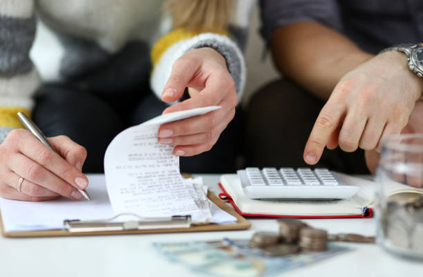 Month revenue of young couple Close-up view of man and woman making account of family income. Writing down and calculating expenses. Attentive review of finance. Calculator on desk. Economy concept cost of living stock pictures, royalty-free photos & images
