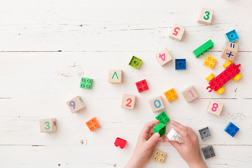 Top view on child's hands playing with wooden cubes with numbers and colorful toy bricks on white wooden background. Baby with toys