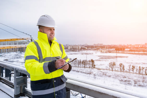 un sorridente costruttore caucasico con un cappello rigido bianco e una giacca fluorescente gialla tiene appunti - inspector built structure engineer clipboard foto e immagini stock