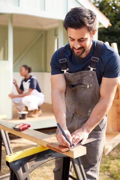 Male Carpenter With Female Apprentice Measuring Wood To Build Outdoor Summerhouse In Garden