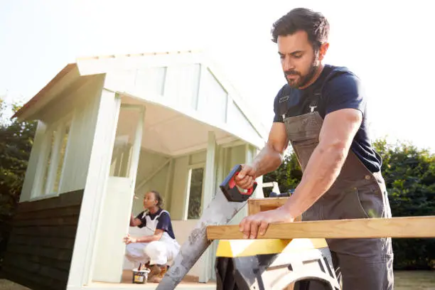 Male Carpenter With Female Apprentice Sawing Wood To Build Outdoor Summerhouse In Garden