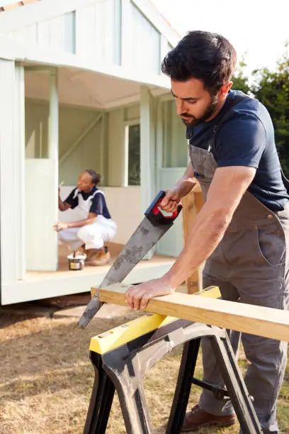 Male Carpenter With Female Apprentice Sawing Wood To Build Outdoor Summerhouse In Garden