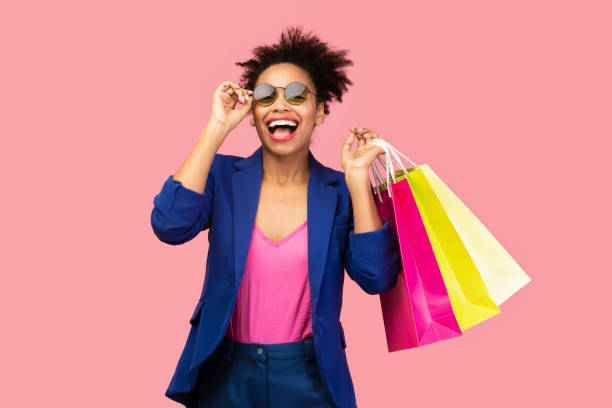 Excited black woman holding shopping bags at pink studio Big Sale. Excited afro girl with shopping bags touching sunglasses, looking at camera over pink studio wall shopaholic stock pictures, royalty-free photos & images