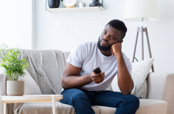 type africain ennuyé s’asseyant à la maison, changeant la tv pour le film - television boredom men sofa photos et images de collection