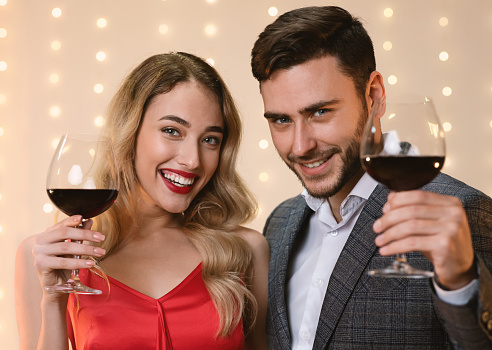 Cheers. Attractive Man And Woman Holding Glasses With Red Wine, Toasting And Smiling At Camera, Celebrating Holidays