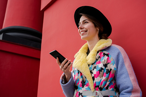 Happy student going to university and searching social networks online. Hipster stylish woman outdoor walking street, using mobile phone, wearing stylish trendy fashionable hat and colorful jacket.