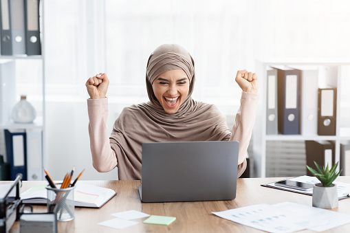 Business Success Concept. Happy Arabic Businesswoman Raising Fists And Exclaming With Joy While Sitting At Laptop In Modern Office, Free Space