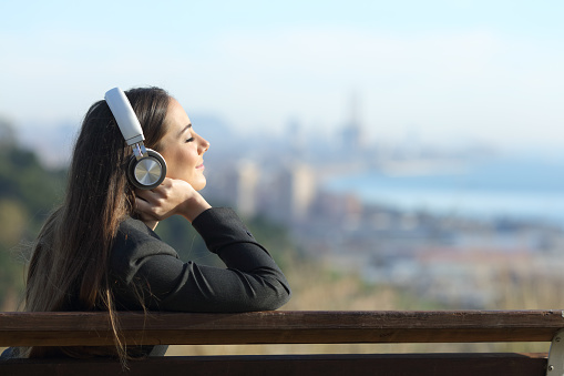 Businesswoman relaxing listening to music wearing headphones sitting on a bench outdoors