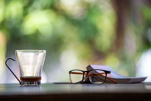 Eyeglasses, Book, Coffee glasses cup with coffee stains stuck on them, then after drinking it on wooden table in bokeh blurred green garden background, Close up shot, Selective focus