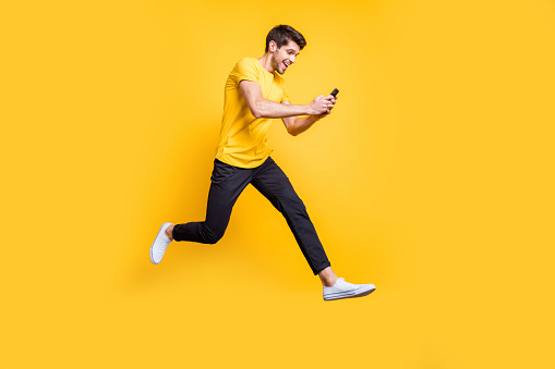 Full body photo of handsome guy jumping high holding telephone, rushing romantic date typing girlfriend sms wear casual t-shirt trousers isolated yellow color background