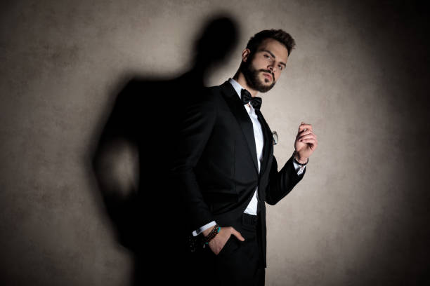 Arrogant groom holding his hand in his pocket and looking over his shoulder while wearing tuxedo and standing on wallpaper studio background