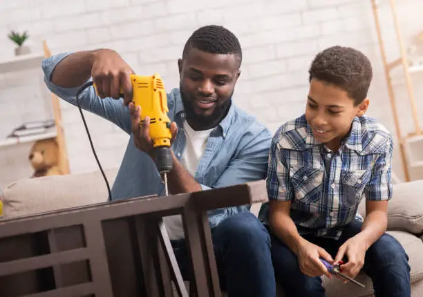 Black father teaching his son how to use drill, perforating chair in living room at home, sharing experience