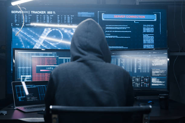 Computer hacker coding on keyboard on a background of monitors. Computer terrorist in a dark hoodie hacking a computer network, types software code on the keyboard and controls a virus attack to hack into government sistems. Digital panels with many open windows with program codes and a windows for loading digital data. computer hacker stock pictures, royalty-free photos & images