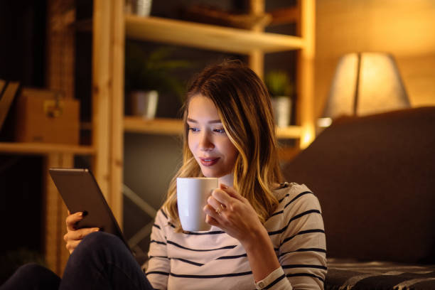 istock Young woman using digital tablet and drinking tea at night in her living room 1205290768