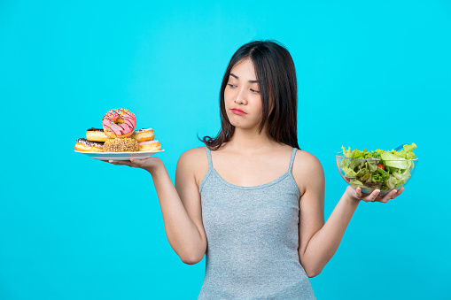 Attractive Asian young woman holding and choosing between disk of donuts or vegetable salad in glasses bowl on isolated blue color background, weight loss and avoid junk food for dieting and HealthyAttractive Asian young woman holding and choosing between disk of donuts or vegetable salad in glasses bowl on isolated blue color background