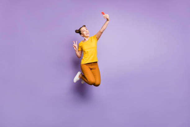 Full length photo of crazy lady jumping high holding telephone taking selfies showing v-sign symbol wear yellow t-shirt pants isolated pastel purple background Full length photo of crazy lady jumping high holding telephone taking, selfies showing v-sign symbol wear yellow t-shirt pants isolated pastel purple background topknot stock pictures, royalty-free photos & images